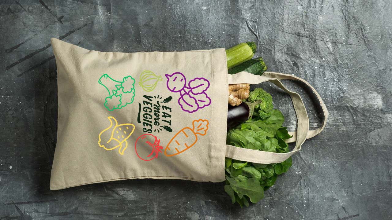 Beige tote bag with colorful vegetable illustrations on a gray surface with groceries.